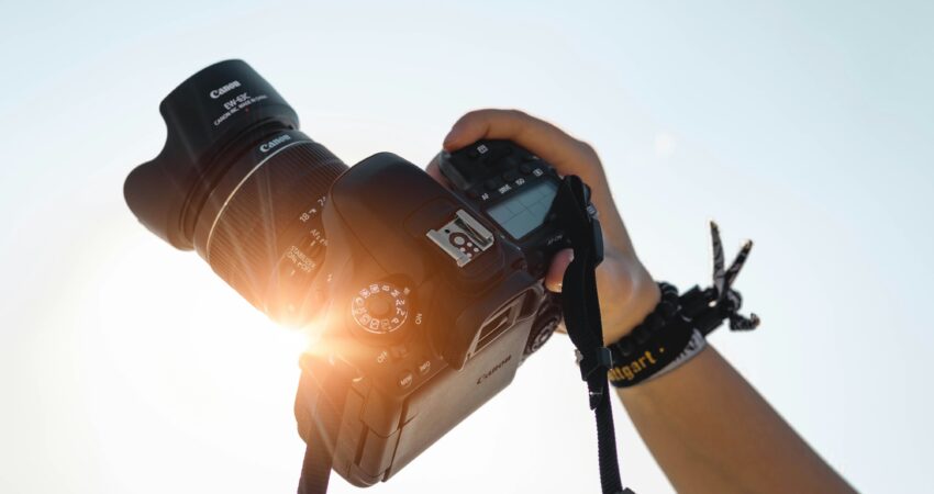 Eine Hand hält eine Canon DSLR-Kamera mit EW-63C-Gegenlichtblende in die Höhe, Sonnenlicht scheint durch das Objektiv