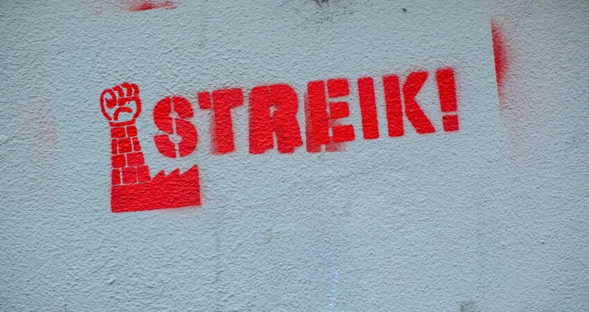 Rotes Graffiti-Schablonenbild an einer Wand mit erhobener Faust über einem Fabriksymbol und dem Schriftzug "Streik!"